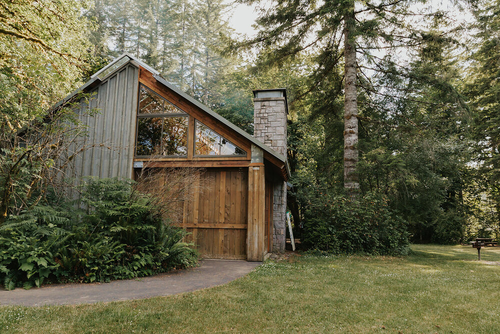 Pictured here is a side view of the Smith Homestead Forest Learning Shelter. The wooden side of the shelter has a moveable wall. There is plentiful trees, grass, ferns, and shrubs that surround the area around the shelter. This can be a great location option for an ADA-accessible elopement.