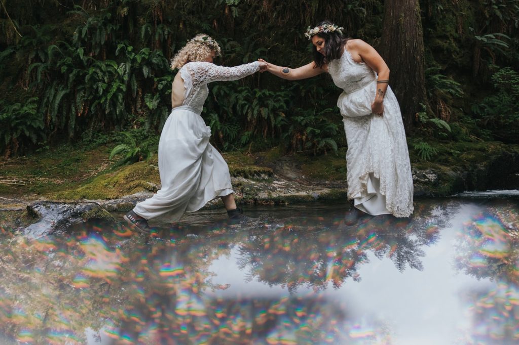 Two women in white wedding dresses are helping each other cross a creek. This is one of the many examples of an affordable elopement dress featured in this blog post!