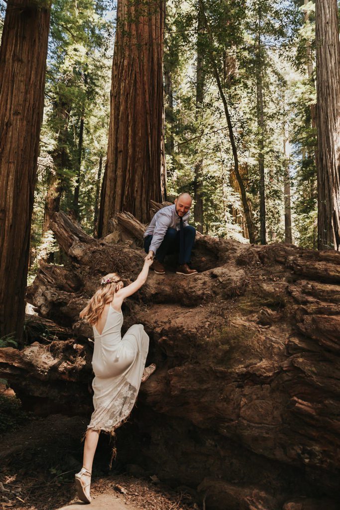 A bride is climbing up a large fallen tree with the help of her partner. This is one of the many examples of an affordable elopement dress featured in this blog post!