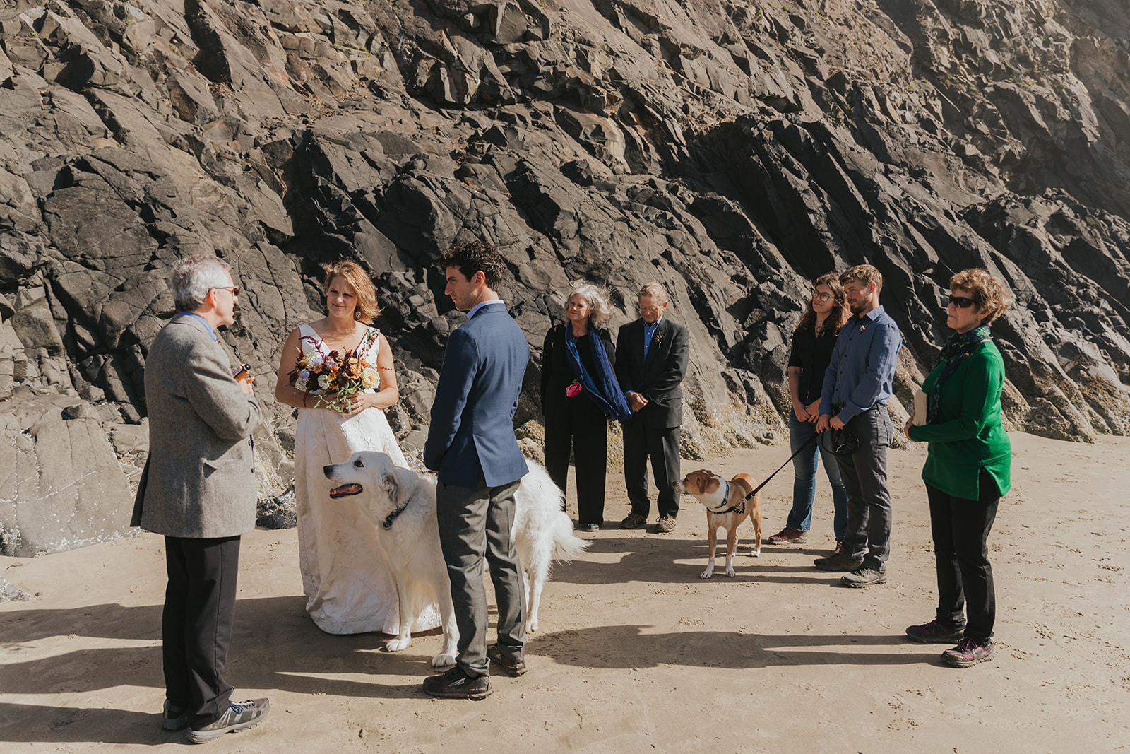 Eloping with family - a couple stands with their dog between them as family watches their elopement ceremony on a rocky beach