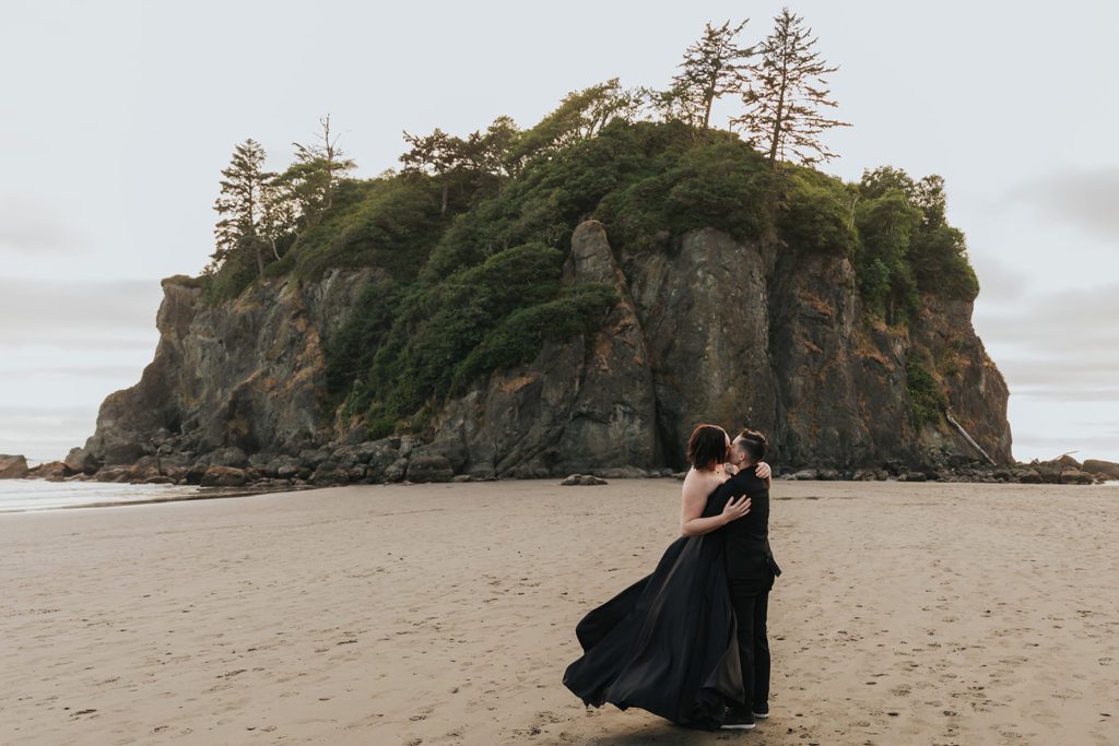 A couple is kissing on the beach while both wearing black.