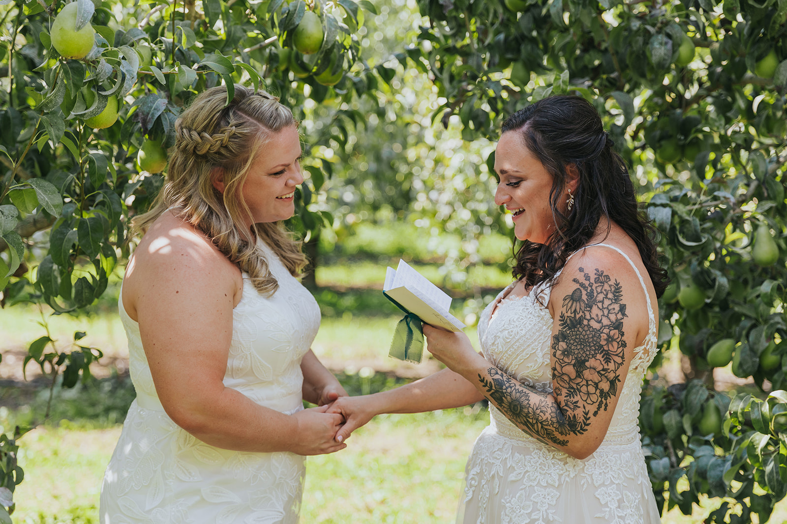 Two brides hold hands in a a pear orchard as they share private vows on their wedding day