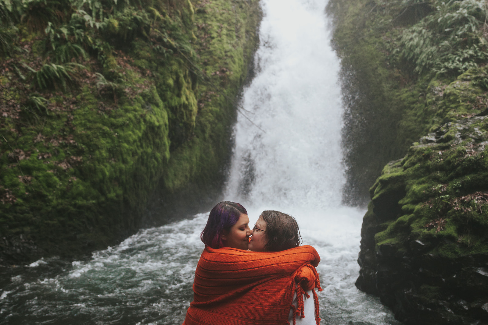 A couple snuggles under a blanket in front of Bridal Veil Falls, a popular engagement photo location near Portland, Oregon.